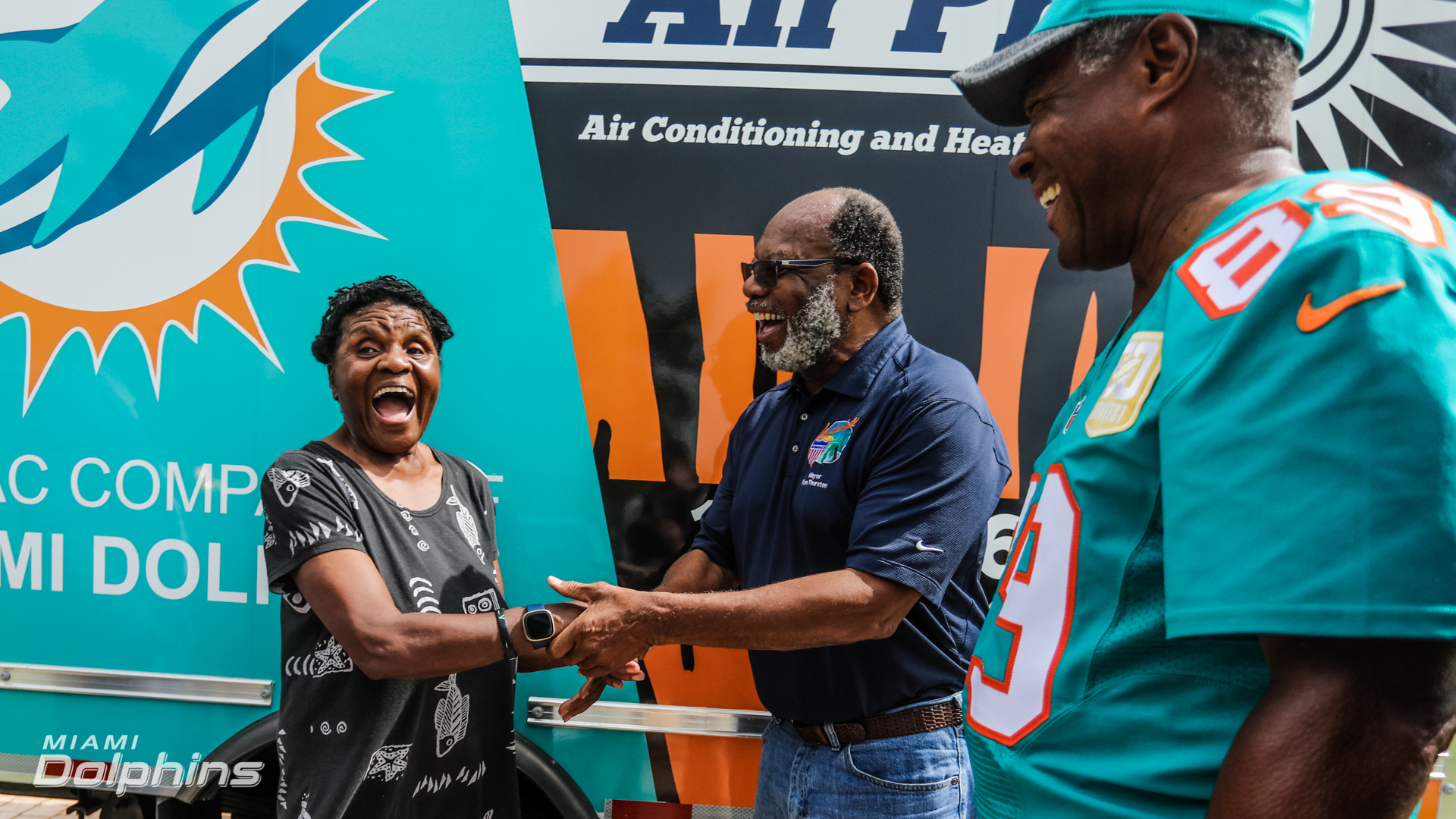 Three people stand smiling and shaking hands in front of a Miami Dolphins-branded vehicle; two wear Dolphins attire, and one has a surprised expression—celebrating with Air Pros USA after receiving a free A/C unit.