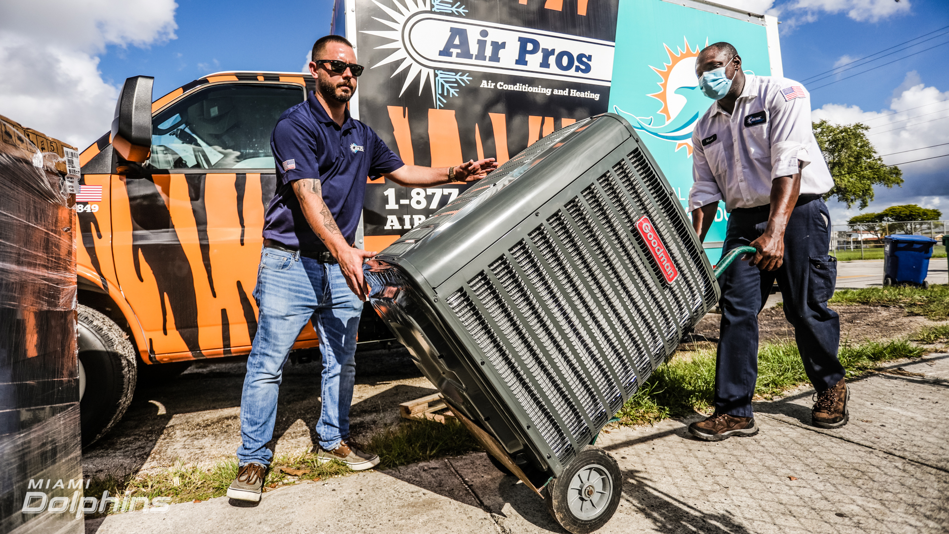 Two workers move a large air conditioning unit on a dolly in front of an Air Pros USA van with a tiger-striped design, offering Miami Dolphins fans a chance to win a Free A/C Unit.