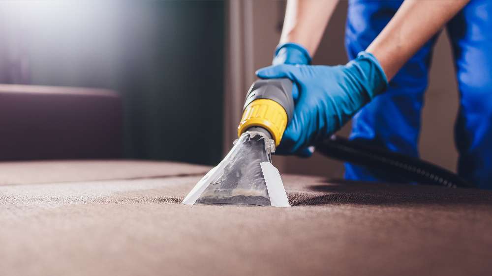 A person wearing blue gloves uses a vacuum cleaner attachment to remove dust and dirt from the surface of a brown upholstered sofa.