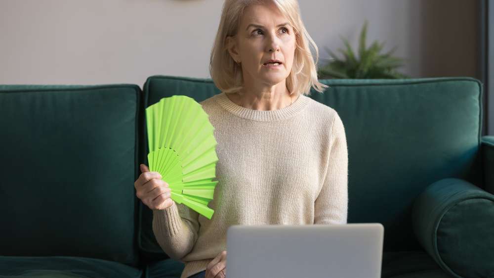 A woman sits on a green couch holding a green fan and looking upward, with an open laptop in front of her, possibly searching for reasons AC not cooling or other air conditioner problems.