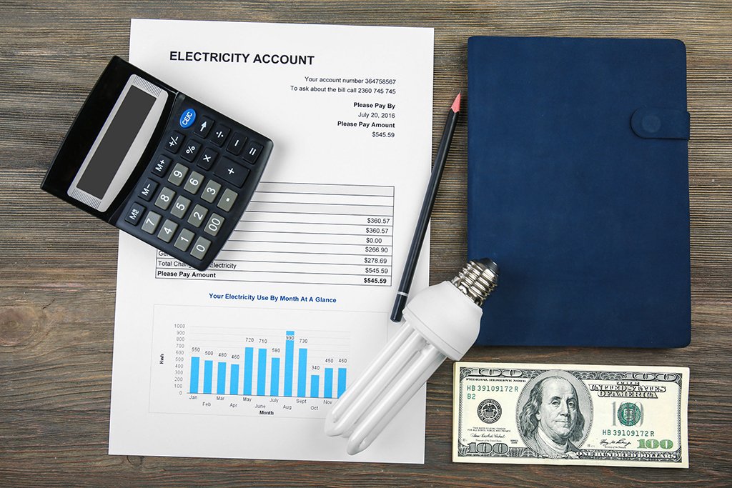 A calculator, pen, notebook, light bulb, $100 bill, and an energy bill with a usage chart are arranged on a wooden surface, offering inspiration for smart summer tips.