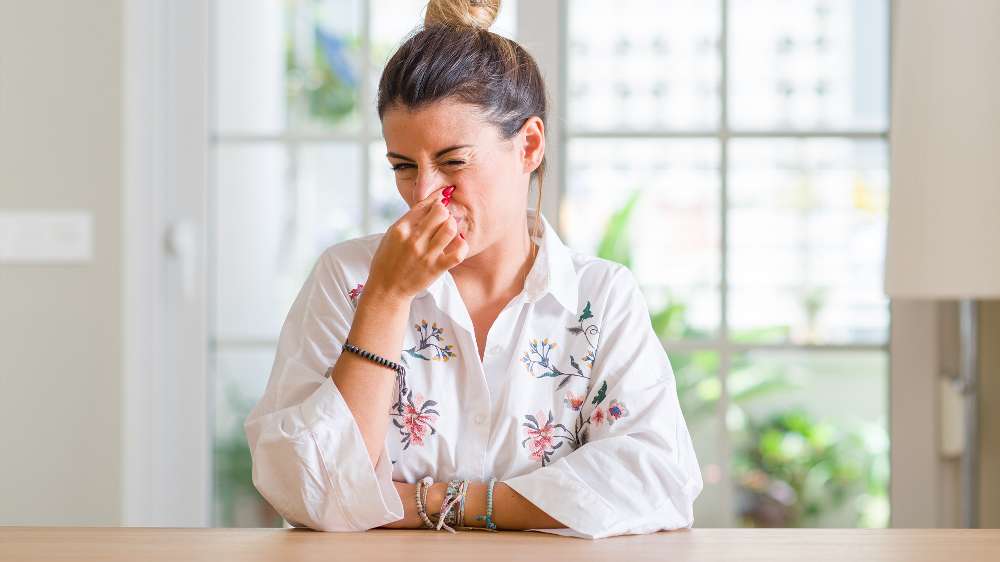 Woman sitting at a table indoors, pinching her nose and making a displeased facial expression, likely reacting to a bad smell—perhaps one of the reasons related to issues with the central AC.