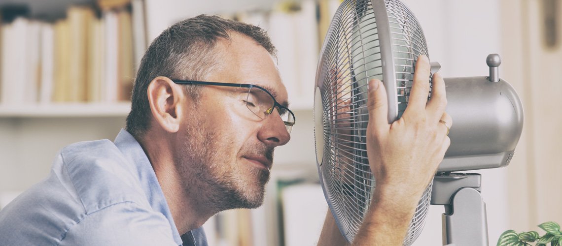 A man with glasses sits close to an electric fan with his eyes closed, appearing to enjoy the cool air—perhaps taking a break from central AC troubleshooting. Bookshelves are visible in the background.