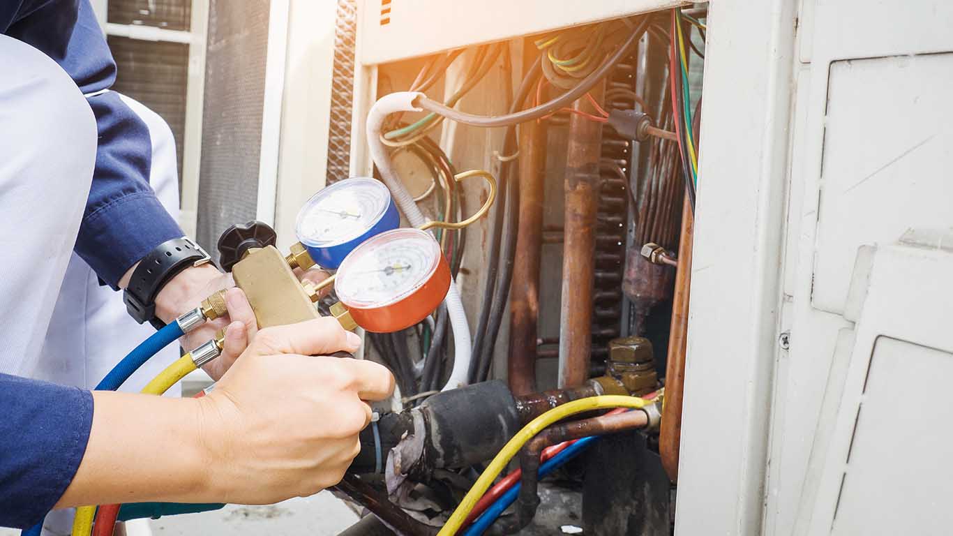 A technician performs air conditioner maintenance, checking an AC unit with manifold gauge hoses and meters attached to pipes and wires as part of a thorough AC checklist.