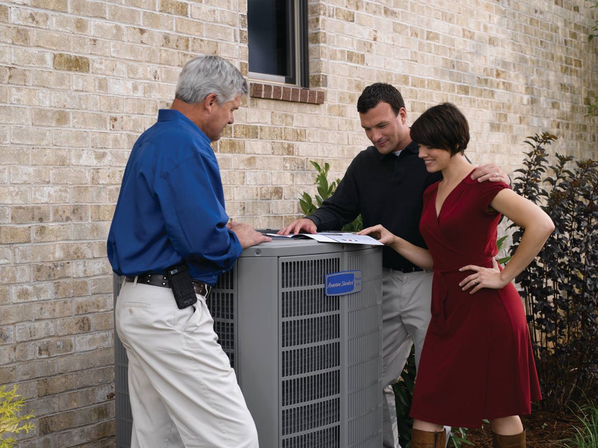 Three people stand outside next to an AC unit by a brick wall, looking at documents on top of the unit—perhaps planning a vacation or discussing travel details.