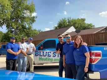 Six people in blue shirts stand in front of two blue pickup trucks with “BLUE STAR Heating and Air” logos, smiling outdoors on a sunny day—ready to share Air Pros tips on Halloween safety, like using glow sticks for better visibility at night.