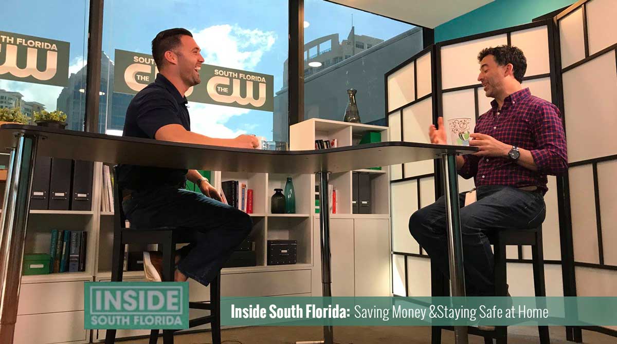 Two men sit at a high TV table on the set of "Inside South Florida," discussing “Saving Money & Staying Safe at Home,” with books, decor, and the Air Pros logo visible in the background.