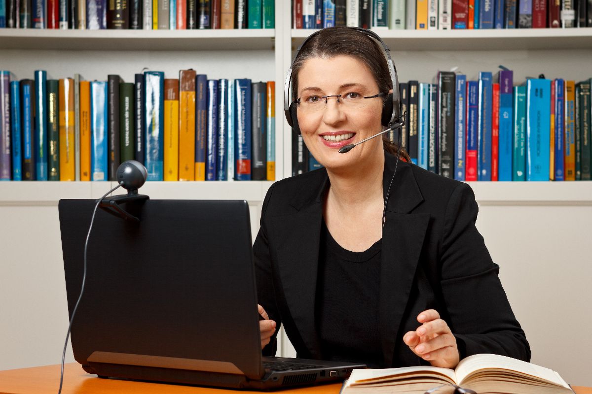 A woman wearing a headset sits at a desk with a laptop, webcam, and open book, smiling in front of bookshelves—an inspiring setting for teachers to connect and engage virtually.