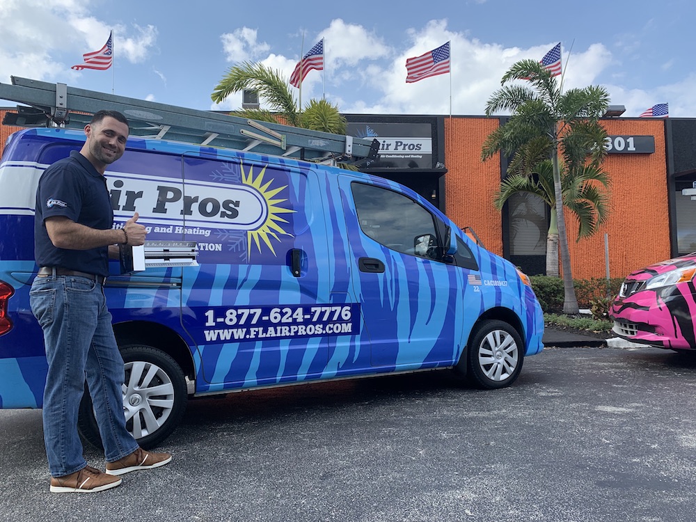 A man stands beside a blue Air Pros USA service van holding a tablet, with an orange building, palm trees, and American flags in the background—a true Power Leader featured in the South Florida Business Journal.