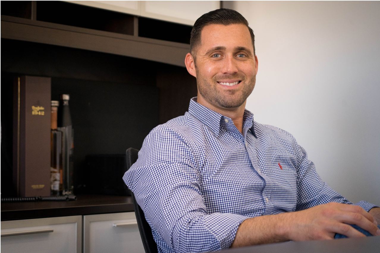 A man with short dark hair and a trimmed beard, wearing a blue checkered shirt, sits and smiles at a desk in an office setting at Air Pros USA, one of South Florida Business Journal’s fastest-growing private companies.