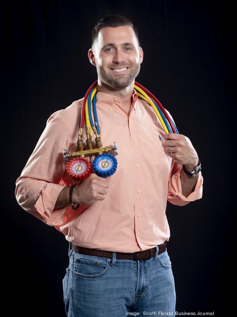 Anthony Perera, founder of Air Pros USA, stands against a black background in a peach button-down and jeans, holding a refrigeration manifold gauge set—a portrait featured in the South Florida Business Journal.