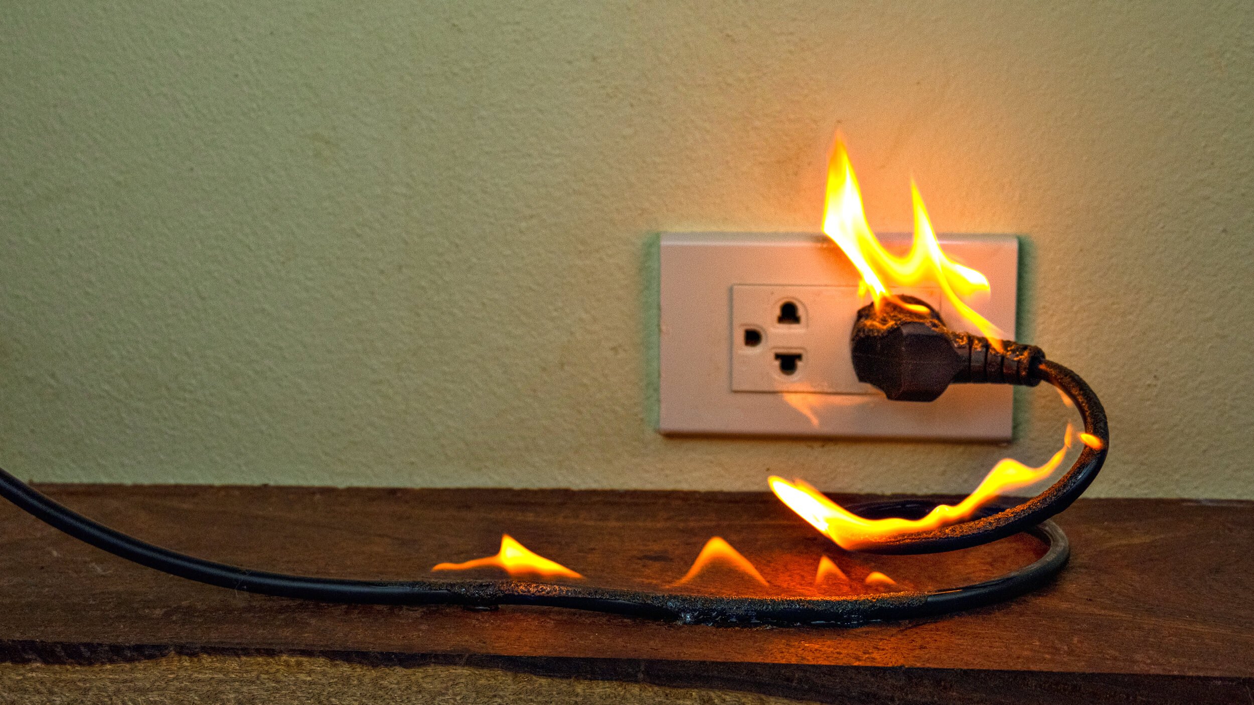 A power cord plugged into a wall outlet is on fire, with visible flames along the length of the cord—a stark reminder of the dangers of electrical problems and the importance of avoiding overloaded outlets.