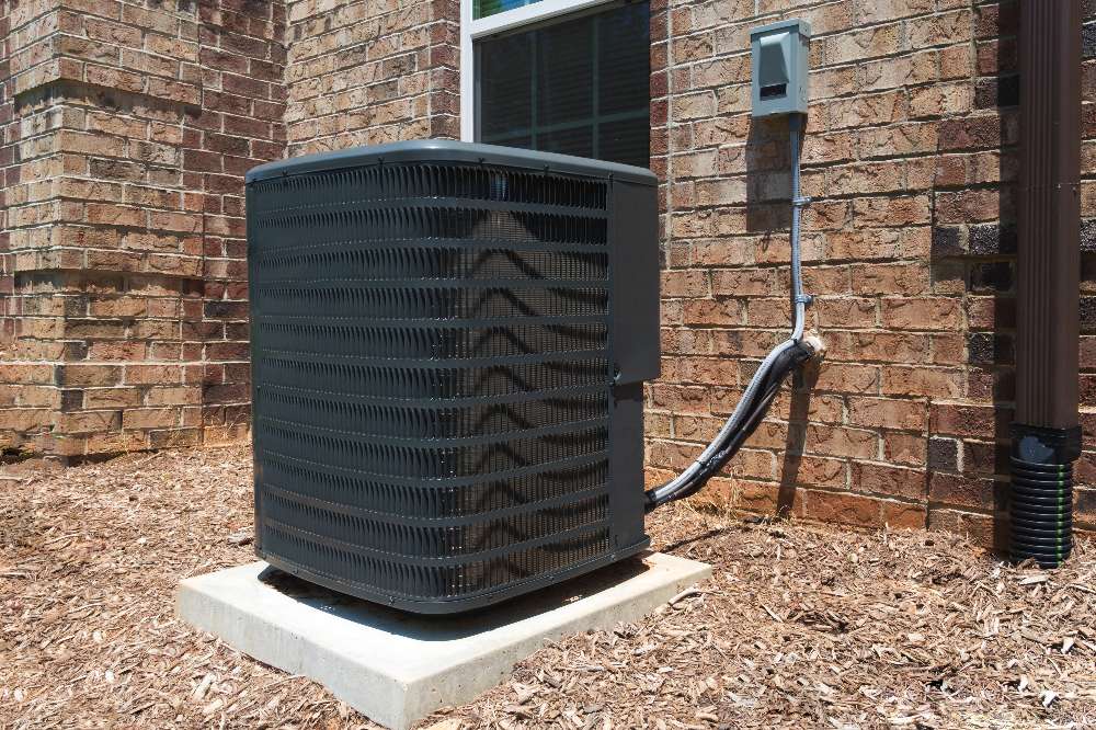 An outdoor air conditioning unit sits on a concrete pad next to a brick house, with electrical and coolant lines connected to the wall—reminding homeowners that regular AC maintenance is essential for peak performance.