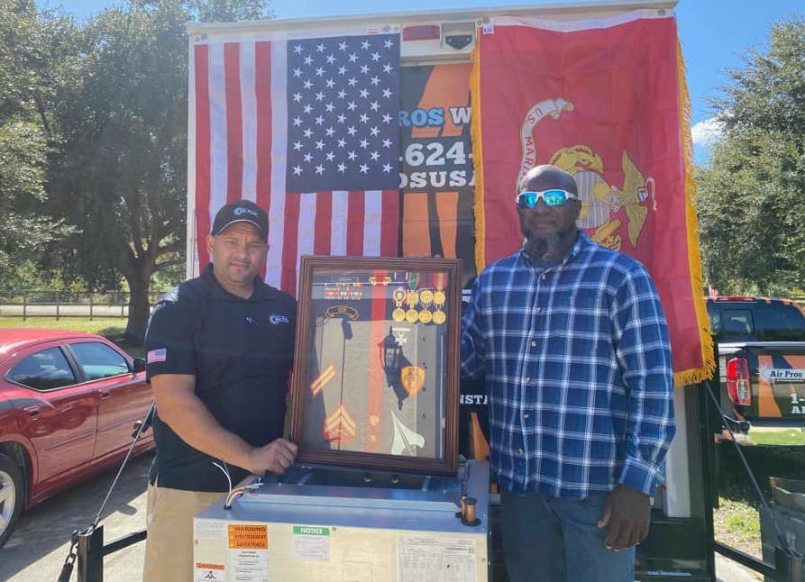 Two men stand in front of American and Marine Corps flags in Fort Myers, holding a framed military memorabilia display outdoors on a sunny day, near an Air Pros air conditioning unit.