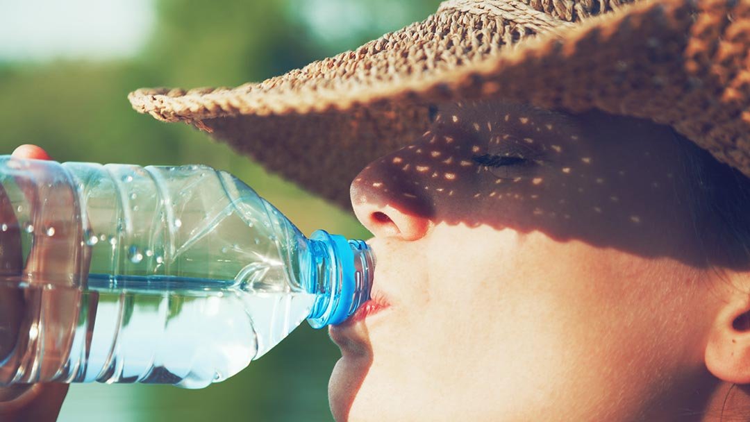 A person wearing a straw hat enjoys cool water from a plastic bottle outdoors in bright sunlight, wishing for some air conditioning to beat the heat.