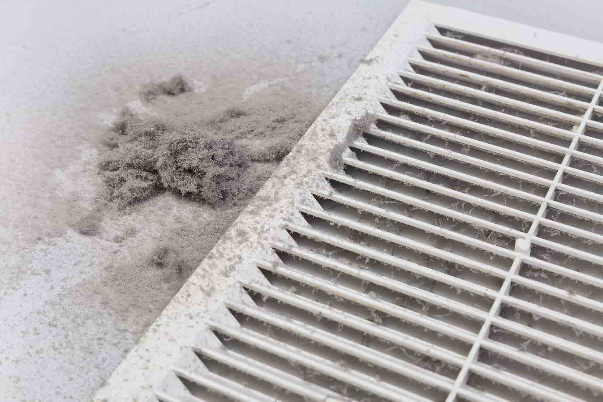 A dirty air vent cover with dust accumulation and a pile of dust beside it on a white surface, showing what can happen if you run an AC without a filter.