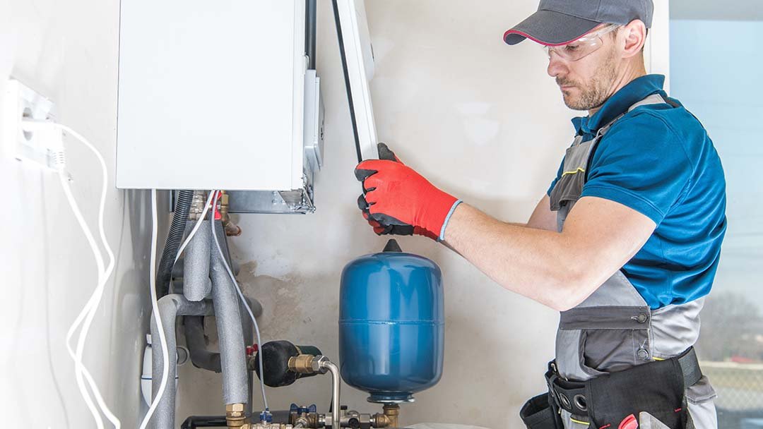 A technician wearing safety glasses and gloves is inspecting or repairing a boiler system, checking for common furnace issues around the pipes and blue pressure tank.
