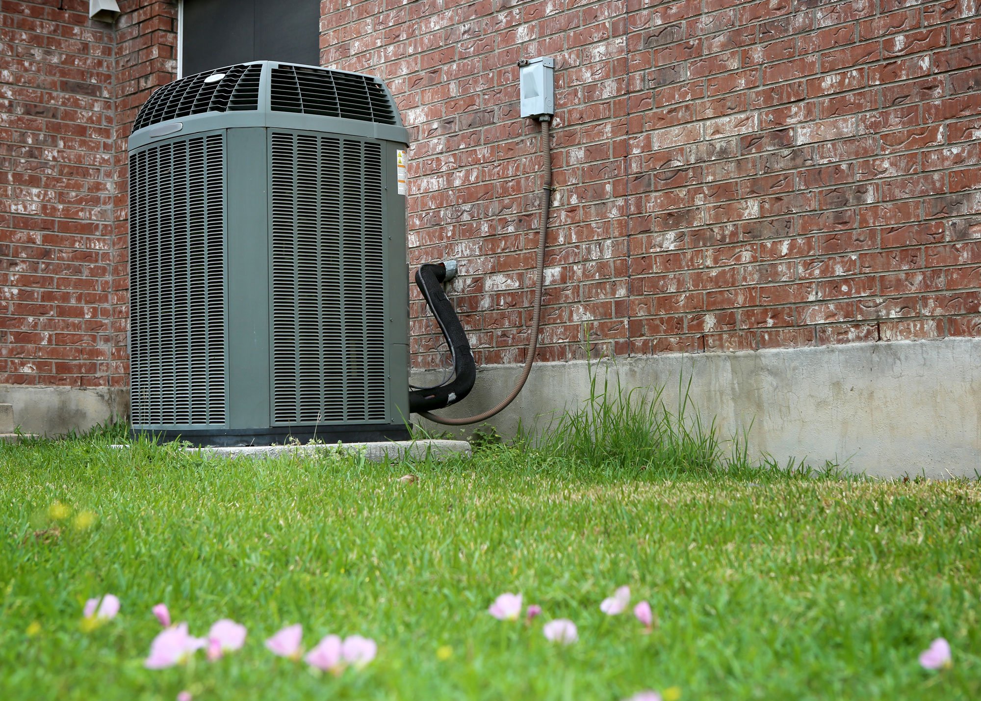 An outdoor air conditioning unit sits next to a brick house with a visible electrical connection—grass and scattered pink petals in the foreground highlight key AC considerations for homeowners.