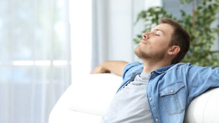 A man sits on a white couch with his arms resting on the back, eyes closed, appearing relaxed—perhaps dealing with common issues of the hottest season. Indoor setting with natural light and green plants in the background.