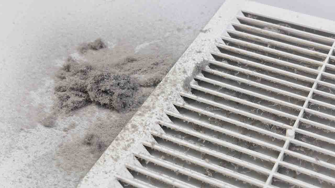 A dusty air vent cover lies on a white surface with a clump of debris nearby, highlighting the importance of central AC maintenance to remove dust from central AC systems.