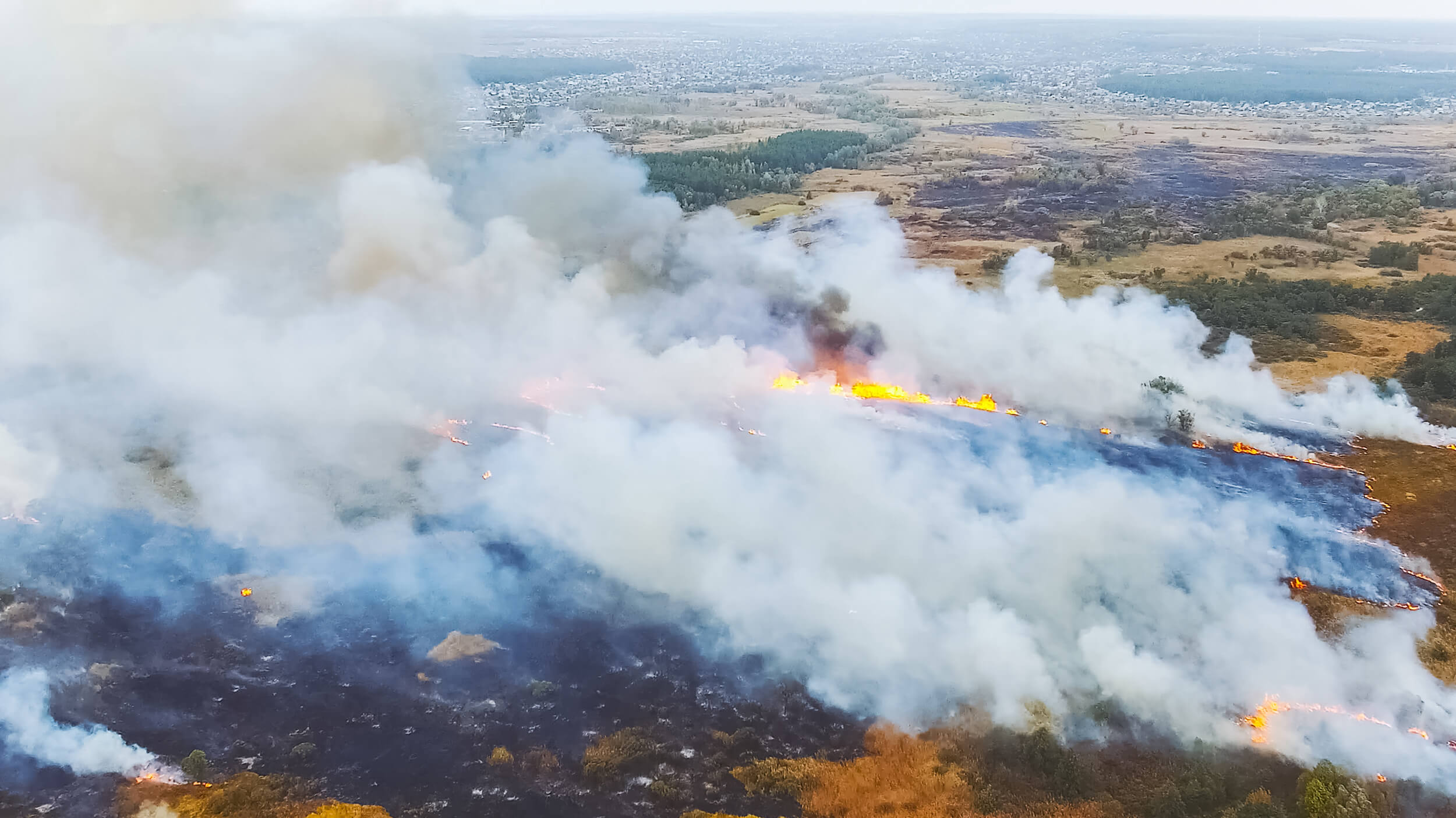Aerial view of a wildfire burning through grasslands and forest, with thick smoke rising—reminding us how wildfires can impact indoor air quality in every home. Patches of unburned land are visible in the distance.