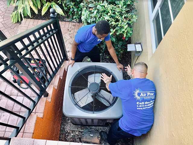 Two technicians in blue shirts perform air conditioner installation beside a building near a staircase and garden, carefully considering AC installation factors for optimal performance.