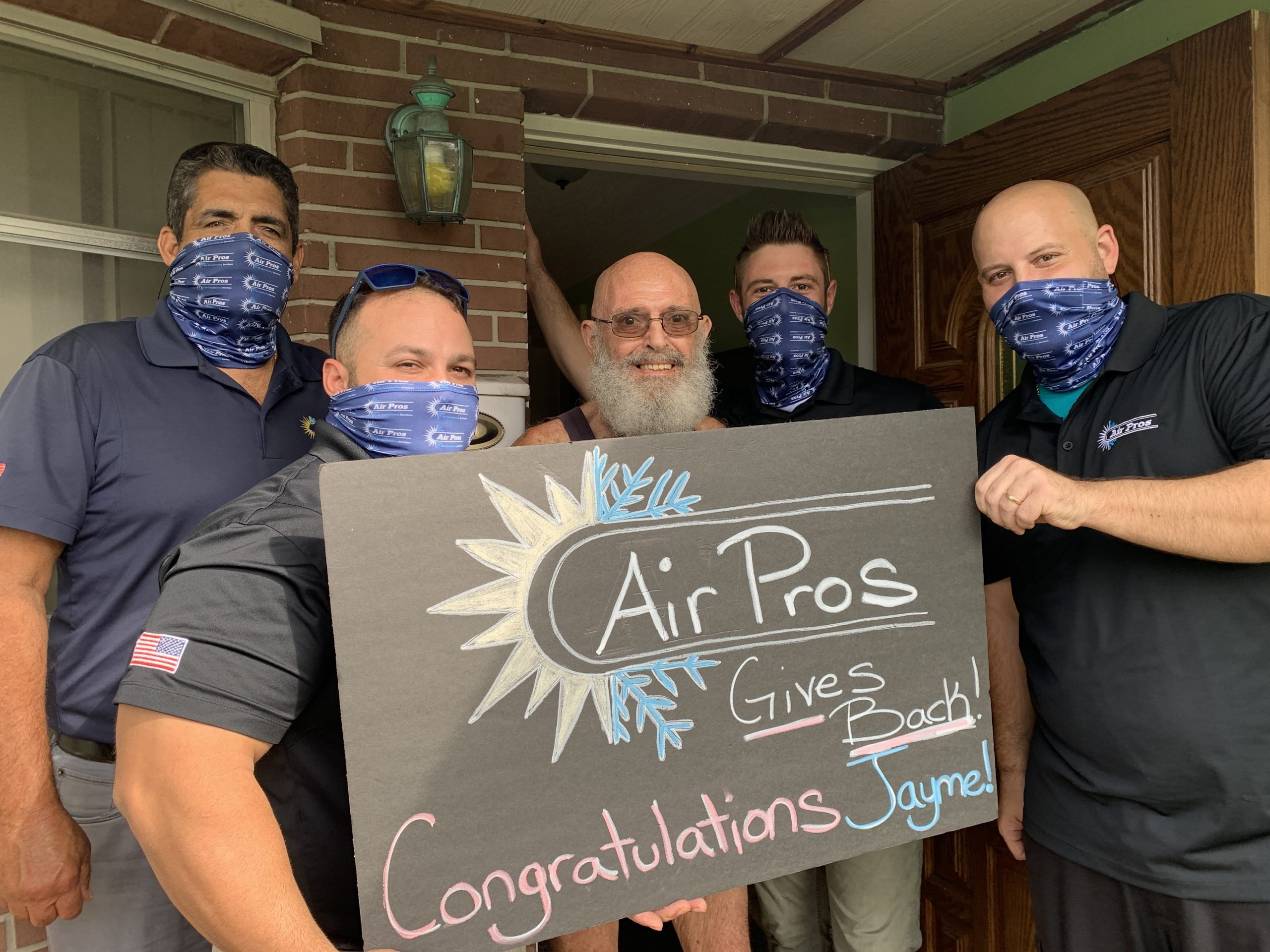 Five men, four in matching Air Pros USA face masks, pose with a sign that reads, “Air Pros Gives Back! Congratulations Jayme Franco!” in front of a house on Veterans Day.