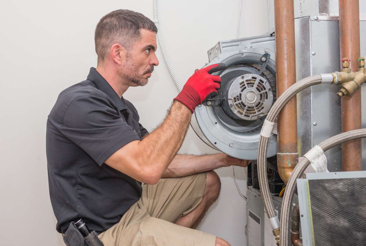 A technician wearing red gloves repairs or inspects the blower motor of an HVAC unit mounted on a wall, addressing potential furnace problems.
