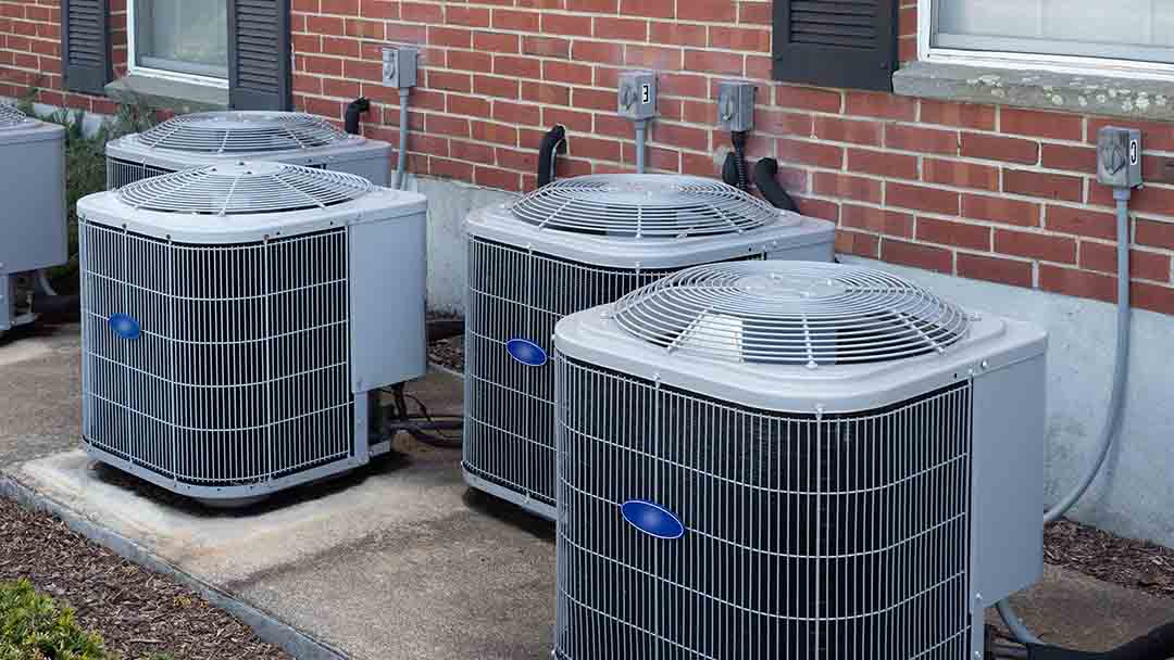 Four central air conditioning units are installed on concrete slabs beside a brick building with several electrical outlets on the wall.