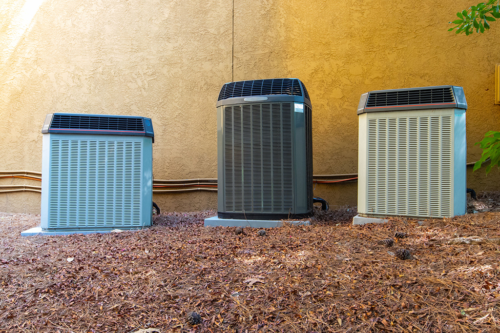 Three outdoor air conditioning units are installed side by side on mulch near a tan stucco wall, with electrical conduit visible behind them—ideal for regular condenser coil maintenance to ensure efficient performance.