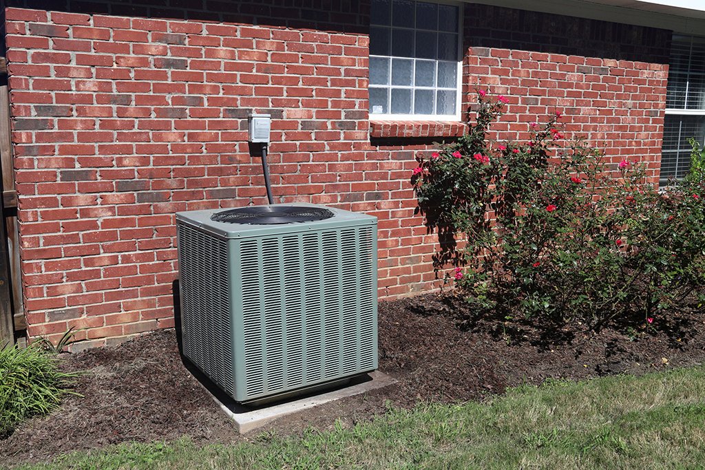 A central air conditioning unit is installed outside a brick house, next to a garden bed with bushes and flowers. Regular AC maintenance ensures the cleanliness of the AC evaporator coils for optimal cooling performance.