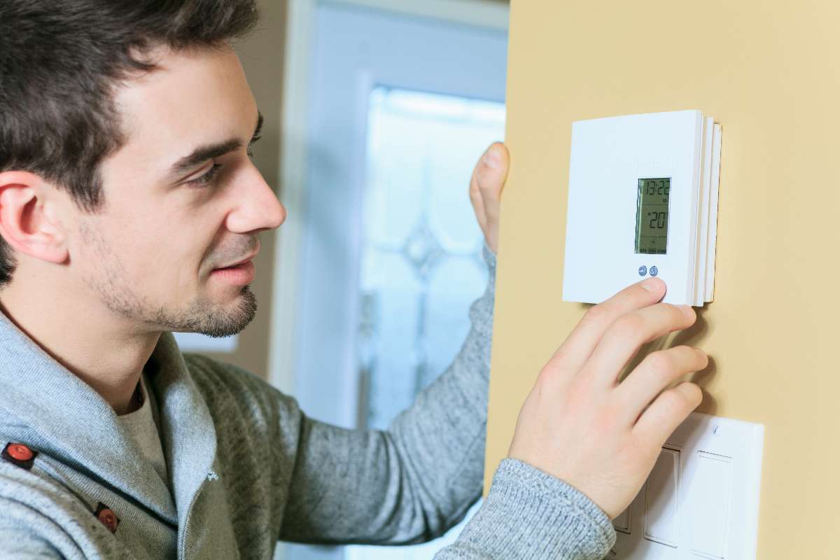 A man adjusts a digital thermostat on a yellow wall, monitoring the temperature display to help maintain optimal indoor air quality.