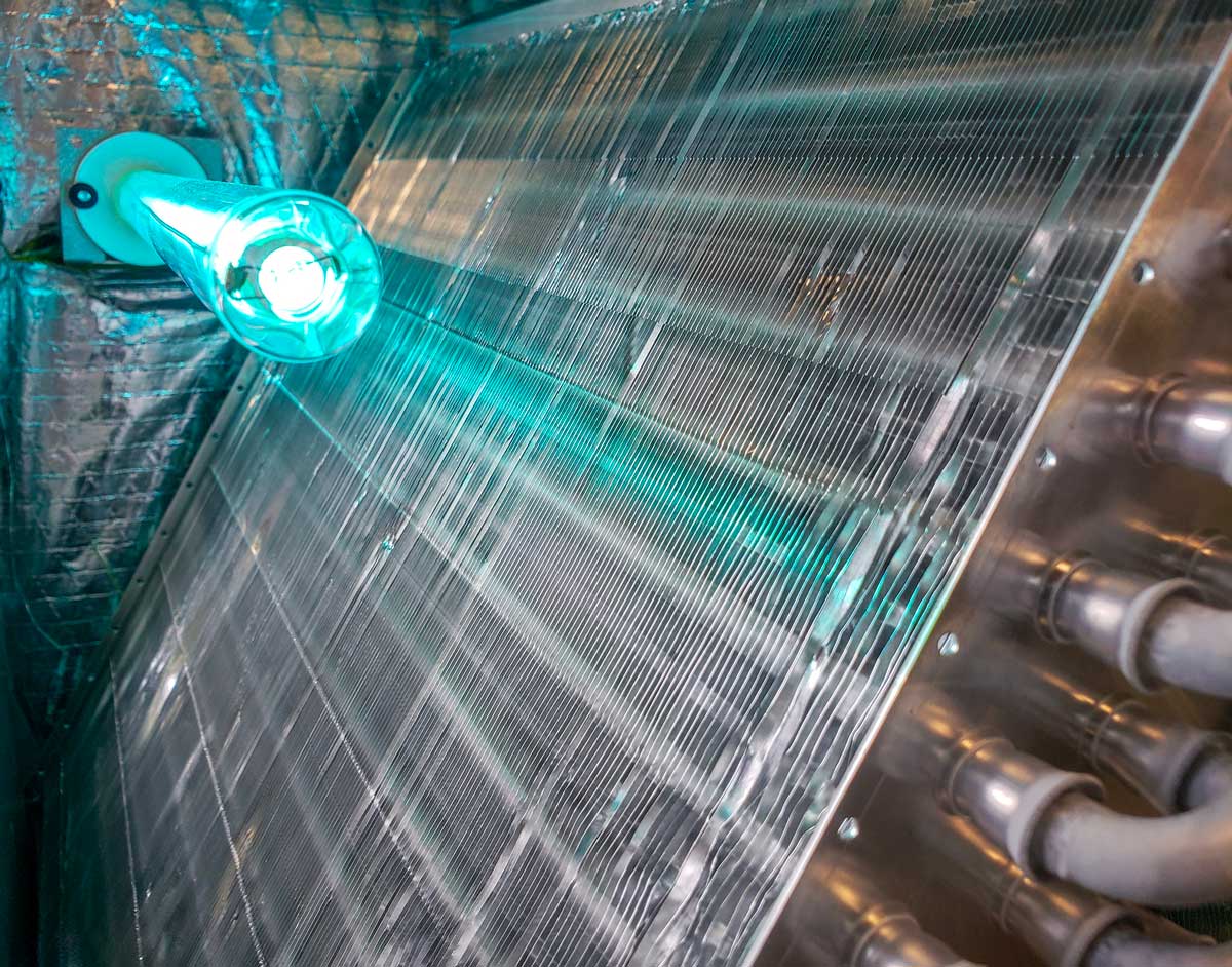 A UV-C air purifier irradiates the surface of a metal heat exchanger with visible fins and attached pipes in an industrial HVAC system.