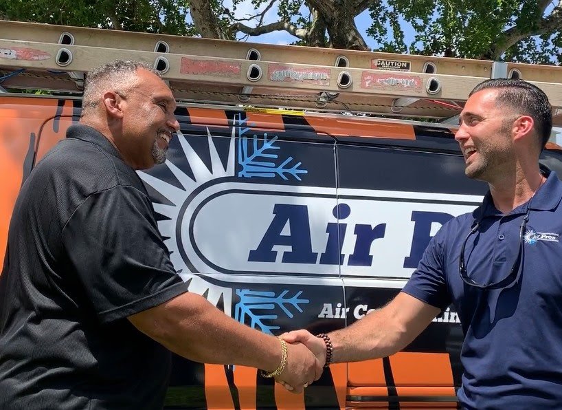Two men, likely HVAC installers, shake hands in front of an "Air Pro" service van with a ladder on top, outdoors on a sunny day.