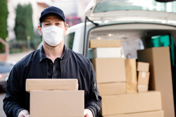 A delivery person wearing a mask and cap holds packages in front of a van filled with cardboard boxes, following COVID-19 policy guidelines.