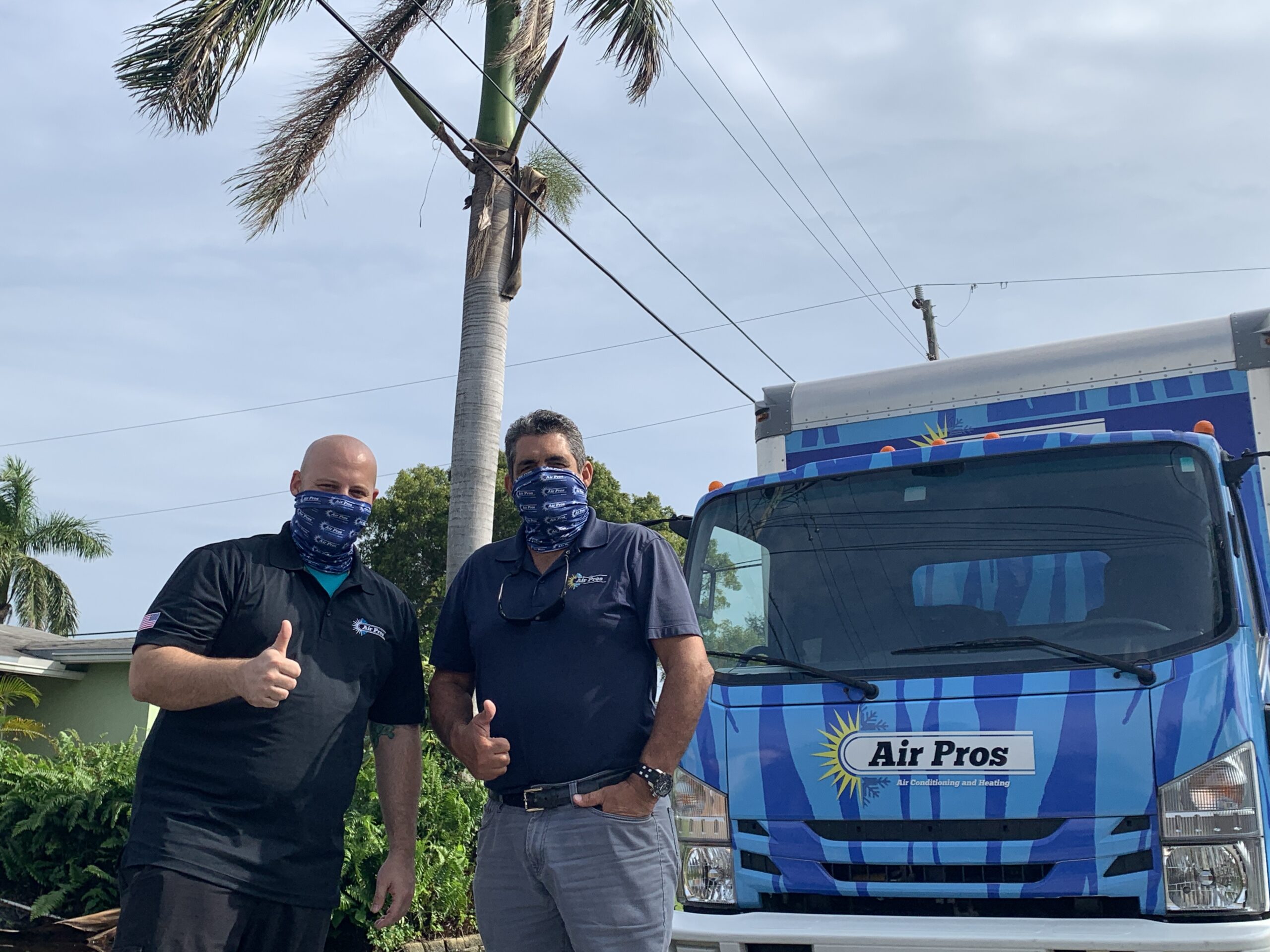 Two men wearing masks and "Air Pros" shirts stand in front of an "Air Pros" service truck, giving thumbs up outside on a sunny day, proudly representing their local AC repair company.