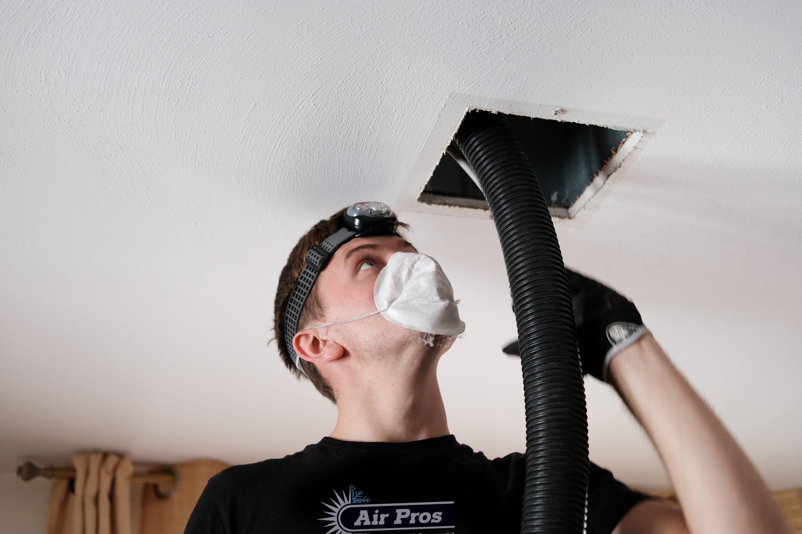 Wearing a mask and headlamp, a person demonstrates the importance of duct cleaning by using a large hose to clean an air duct in the ceiling—an essential task, especially before summer.