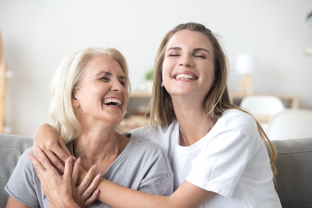 Two women sit on a couch, smiling and hugging each other, looking happy and relaxed in a bright room with improved home air quality thanks to HVAC UV lights.