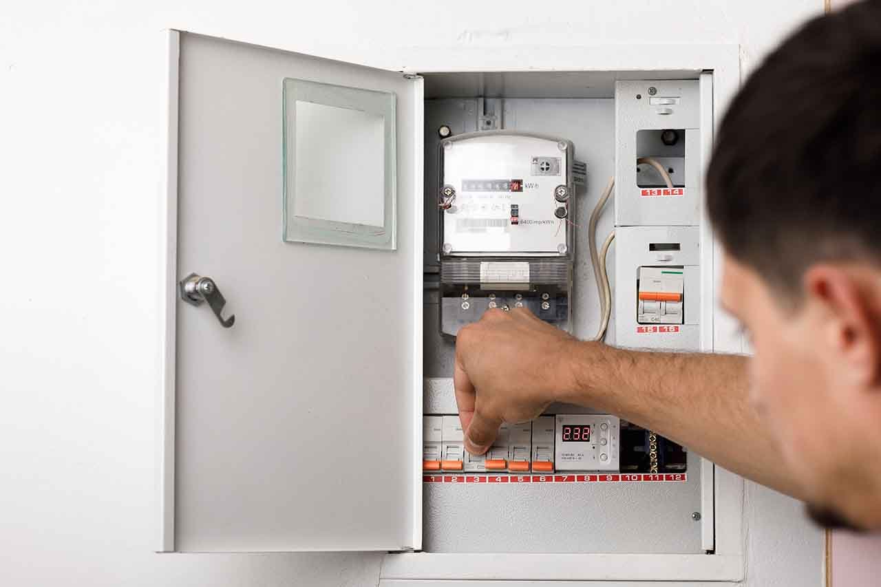 A person adjusts switches inside an open electrical panel mounted on a white wall, checking for a circuit breaker issue that could be causing the air conditioner to trip.