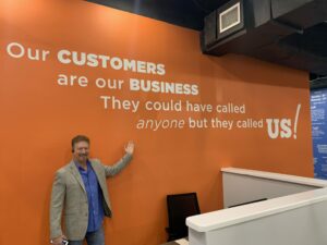 A man in a blazer stands in front of an orange wall with large white text about customer service, representing the Air Pros leadership team as he gestures towards the text.