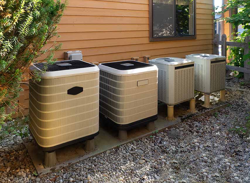 Four outdoor air conditioning units are lined up on a concrete pad next to a house with wood siding and a nearby shrub, where blocked airflow from plants is one of the common AC compressor causes that can lead to AC failure.