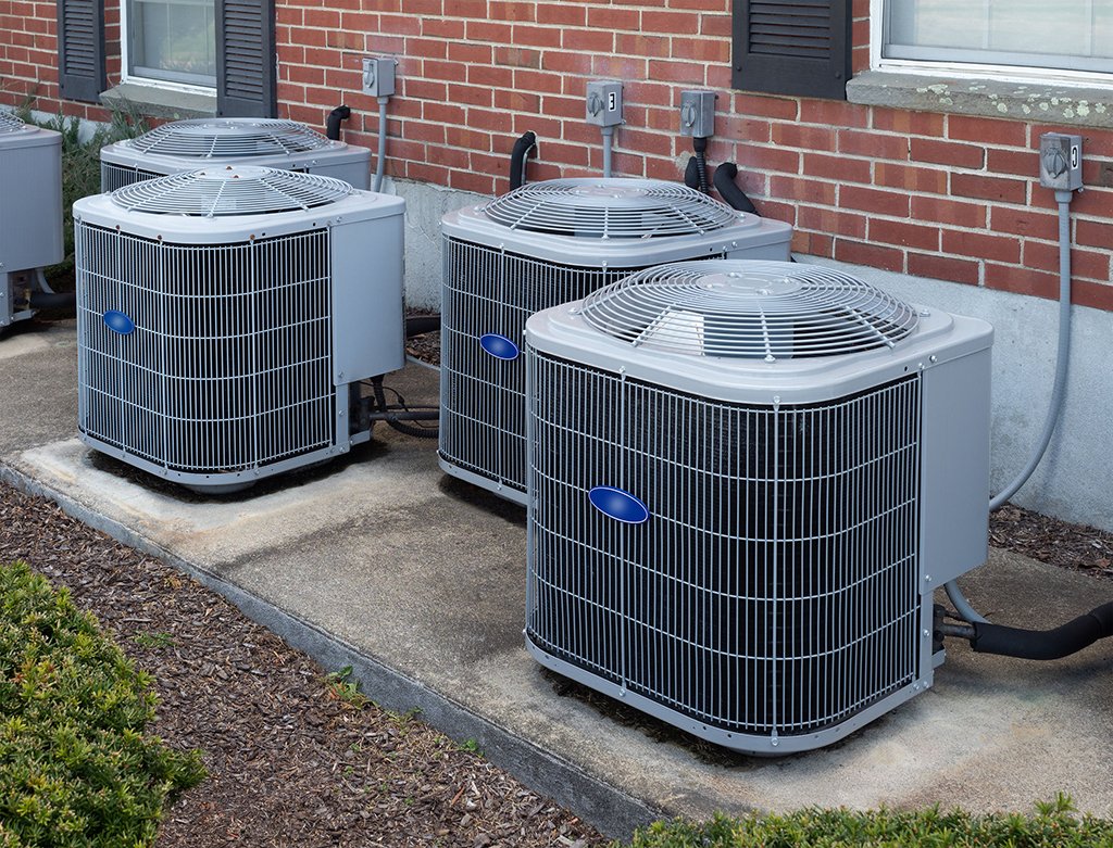 Three outdoor air conditioning units are placed on concrete pads next to a brick building, with electrical connections and a visible condensate drain line behind them—a reminder of the importance of regular air conditioner maintenance.