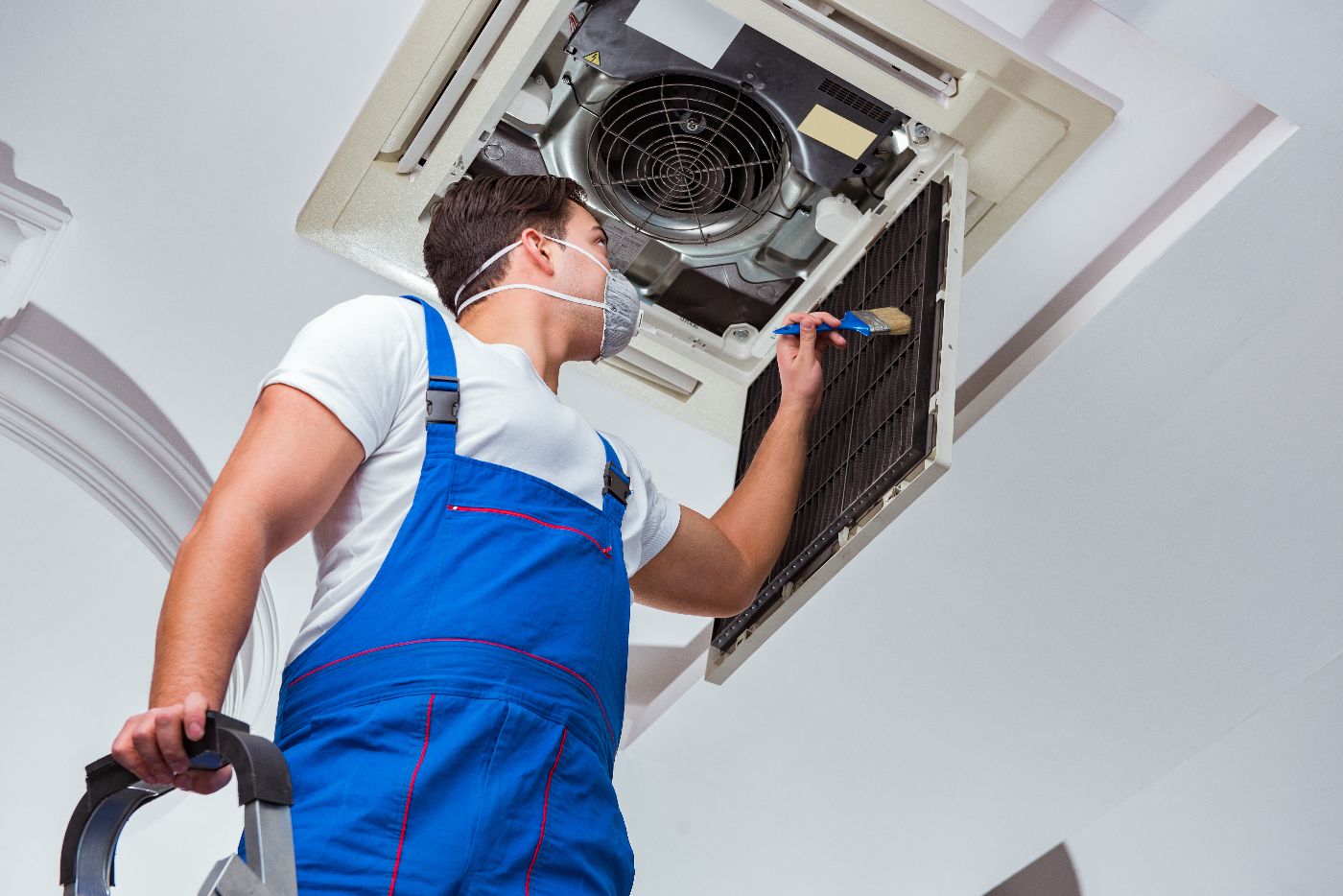 A worker in blue overalls and a face mask cleans an air conditioning vent on the ceiling while standing on a step ladder, ensuring proper air filter maintenance for optimal performance.