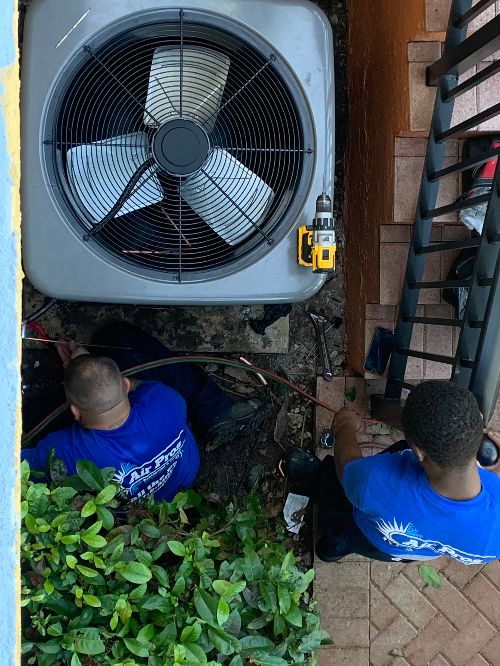 Two workers in blue shirts are repairing or installing an outdoor AC unit next to a staircase, keeping it in tip-top shape for summer, with tools and cables visible around them.