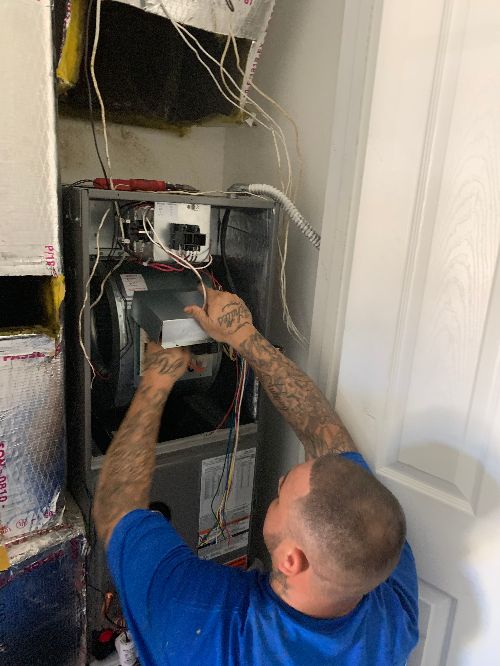 A person in a blue shirt performs AC maintenance, repairing or installing wiring inside an open HVAC unit with exposed wires and ductwork visible.