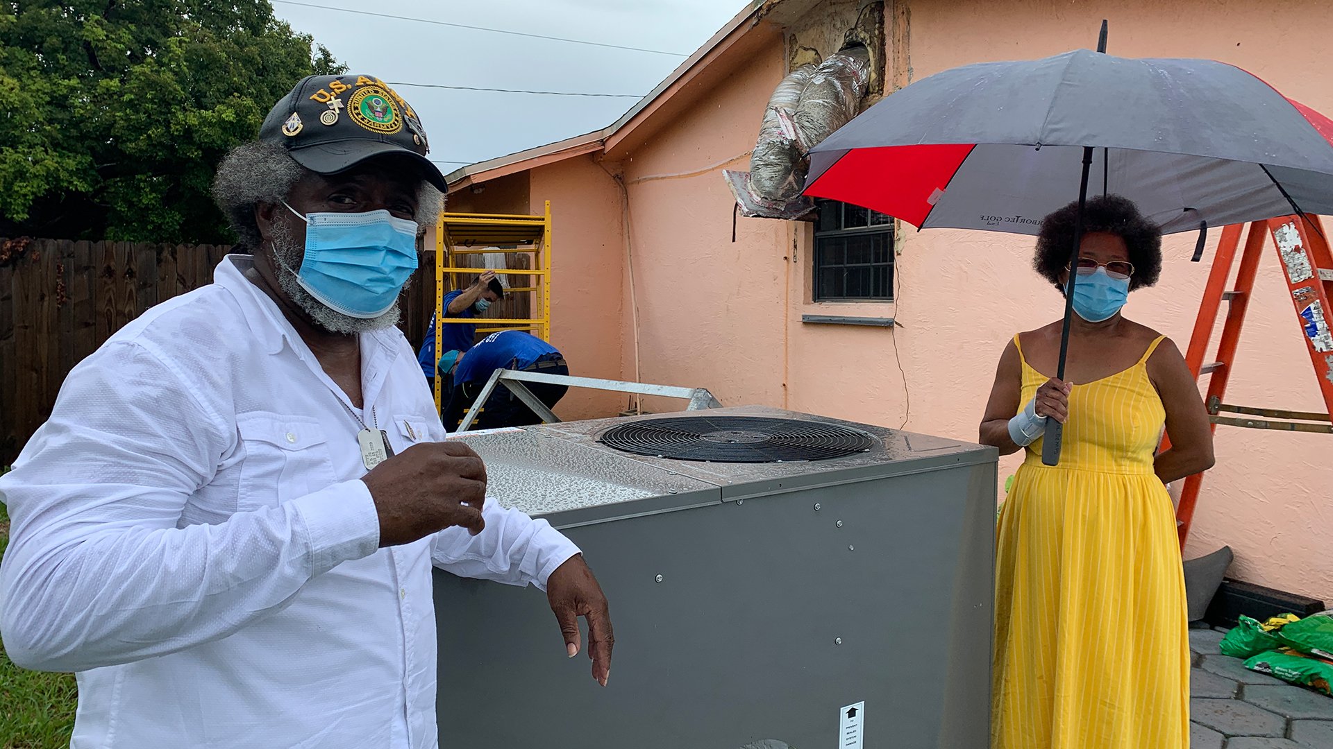 Two people wearing masks stand beside a Goulds Florida air-conditioning unit; one holds an umbrella and the other, in a cap, stands nearby. A house and ladder are visible in the background.