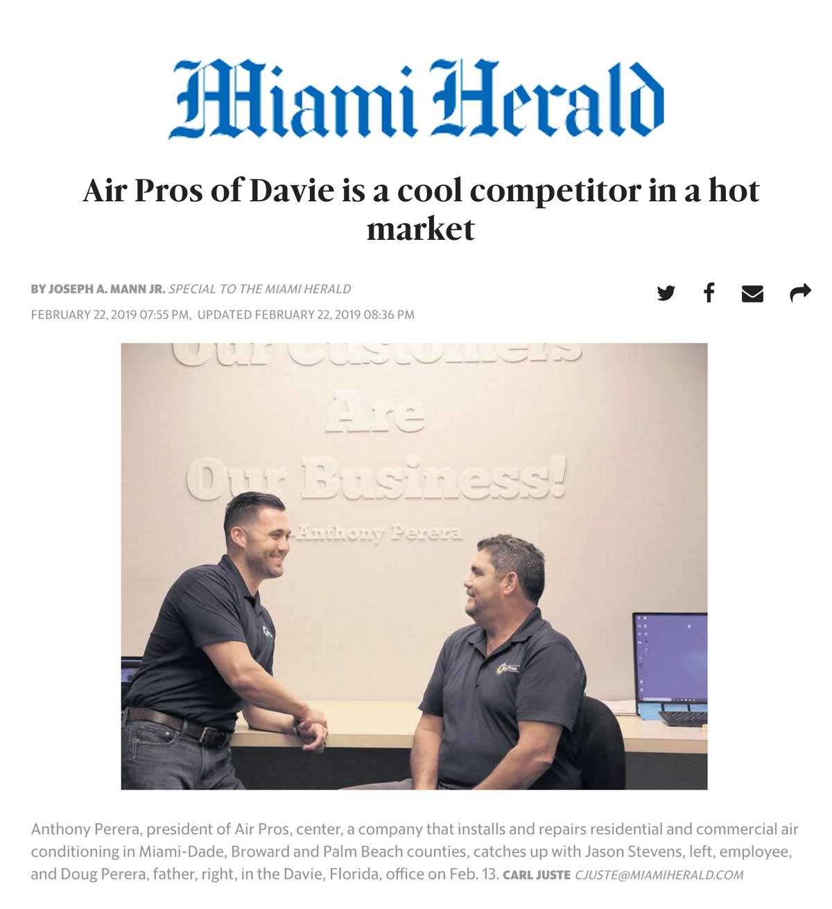 Two men sit at a desk in an HVAC Business office with a sign reading "Our Customers Are Our Business!" on the wall behind them. A computer monitor is visible on the right, highlighting their Air Pros feature in the Miami Herald.