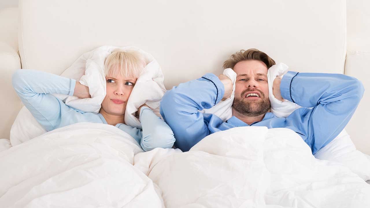 A man and woman in bed cover their ears with pillows, both appearing frustrated or annoyed by central AC noise.