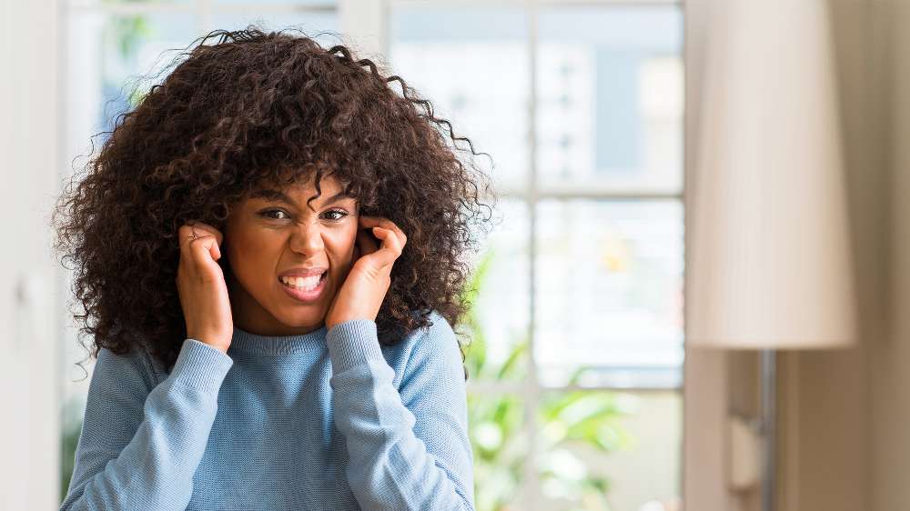 A person in a blue sweater covers their ears with their fingers and makes a frustrated facial expression indoors, likely reacting to loud Central AC noises.