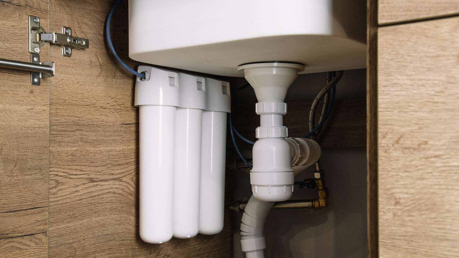 Three white water filtration cartridges and plumbing pipes are installed under a sink inside a wooden cabinet, ensuring clean water flows into your home.